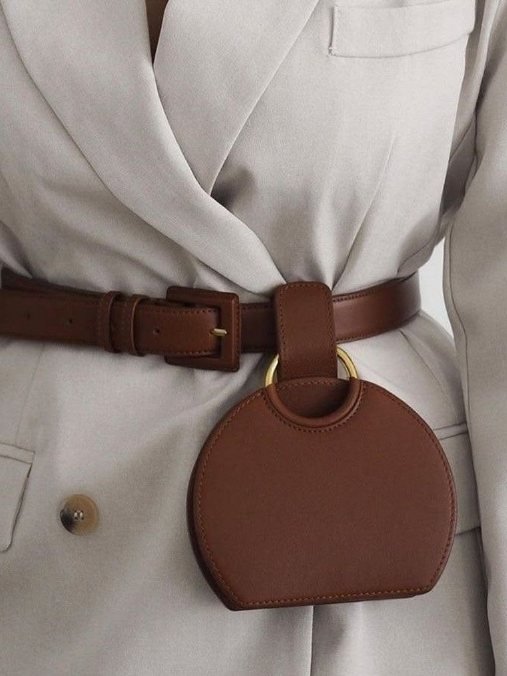 A brown leather DK Luxora belt with a round pouch, worn over a cream blazer
